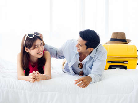 Happy couple travelers on honeymoon trip. Asian man in denim shirt and young beautiful woman smile and lying together with yellow suitcases on bed in bedroom. Happy holiday. Colorful summer vacation.の写真素材