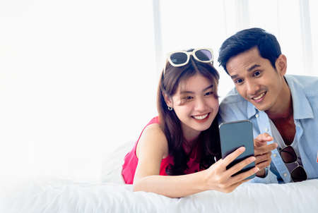Happy Asian couple holds mobile phone. Young man and woman in casual feel happy with smiling and excited while having and looking at smartphone screen while lying on bed in white room with copy space.の写真素材