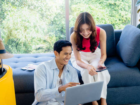Happy Asian couple, young woman and man looking and using laptop computer and smartphone together for flight booking and trip information in living room near luggage, travel holiday summer vacation.の写真素材