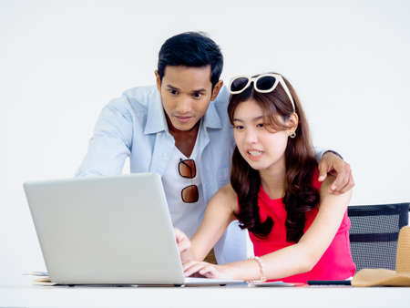 Happy Asian couple, young woman and man using laptop computer on desk together for flight booking, hotel room and trip information in office, ready to travel, happy holiday concept.の写真素材