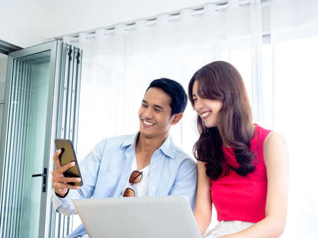 Happy Asian couple, young man and woman in casual looking at smart mobile phone screen while using and working with laptop computer together, video call with their friends in bright room.の写真素材
