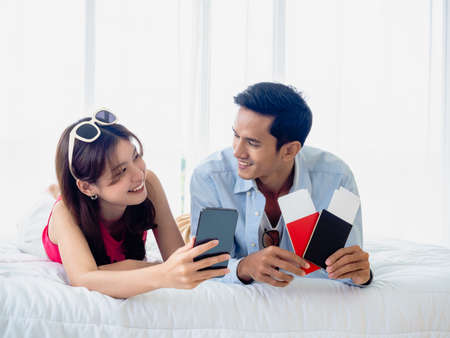 Happy Asian couple with flight booking for summer trip, travel abroad. Young man and woman in casual holding smartphone and two passports and tickets while lying in bed together, look at each other.の写真素材