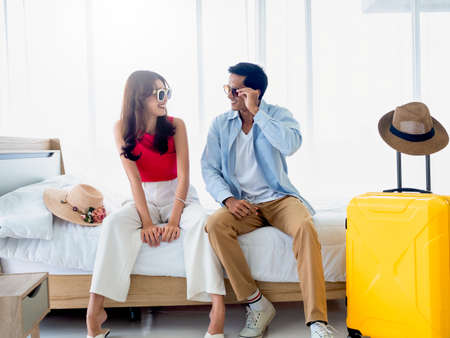 Happy Asian couple in summer, young man and woman wearing sunglasses with smile, sitting on the bed in the bright bedroom near yellow luggage and beach hat, holiday travel, summer vacation concept.の写真素材