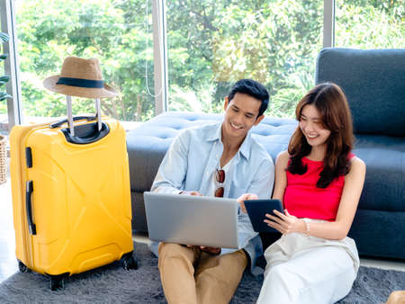 Happy Asian couple, young man and woman looking and using laptop computer and smartphone together for flight booking and trip information in living room near luggage, travel holiday summer vacation.の写真素材