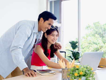 Asian couple, young woman and man looking at laptop computer screen on desk together for travel booking, flight, hotel room and trip information in office, happy summer holiday concept.の写真素材