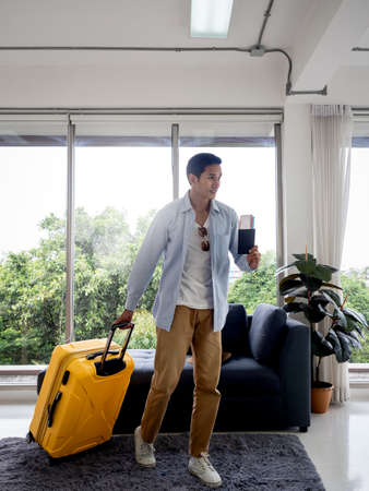 A man with yellow luggage. Handsome Asian male in denim shirt walking while holding passport near the glass window, vertical style. Summer holiday travel vacation concept. Time to journey.の写真素材