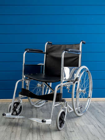 Empty steel wheelchair with black leather seat and backrest on the blue wooden plank wall background, vertical style. Chair with wheels for use as transport by person who unable walk, illness, injury.の写真素材