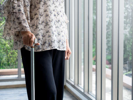 Close up senior woman's hand holding a walking cane standing alone near the glass window in the room with copy space. Lonely elder female, the grandmother with a walking stick.の写真素材