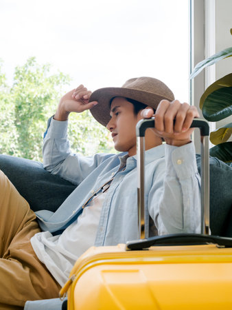 A man with yellow luggage. Handsome Asian male in denim shirt sitting and waiting and thinking while holding suitcase near the glass window, vertical style. Summer holiday travel vacation concept.の写真素材
