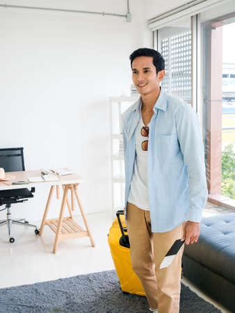 A man with yellow luggage. Handsome Asian male in denim shirt walking while holding passport near the glass window in office, vertical style. Summer holiday travel vacation concept. Time to journey.の写真素材