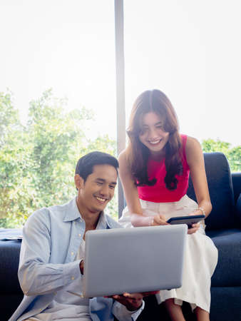 Happy Asian couple, young woman sit on sofa and man on floor looking and using laptop computer and...