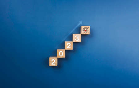 2023 year numbers on on wooden blocks stack, chart steps with shining rise up arrow to the target icon on blue background, business growth process to success, and economic improvement concept of 2023.の写真素材