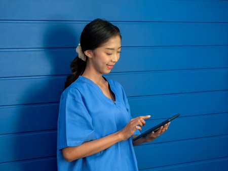 Smiling Asian female doctor or nurse in blue scrubs suit using smart mobile phone while standing on blue background, looking at screen, working online, chat, consulting patient, telemedicine concept.の写真素材