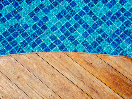 Top view of empty wooden plank table or deck floor curved shape in front of the blurred background of blue mosaic tiles grid pattern in swimming pool. Empty space on poolside, summer background.の写真素材