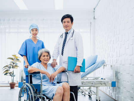 Male surgeon doctors and professional female nursing assistants take care senior woman patients in wheelchair near the bed in white clean recovering room in hospital. Health care and medical concept.の写真素材