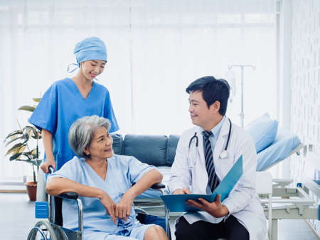 Happy senior woman patients in wheelchair smiling with male surgeon doctors and professional female nursing assistants in white recovering room in hospital. Health care and medical concept.の写真素材