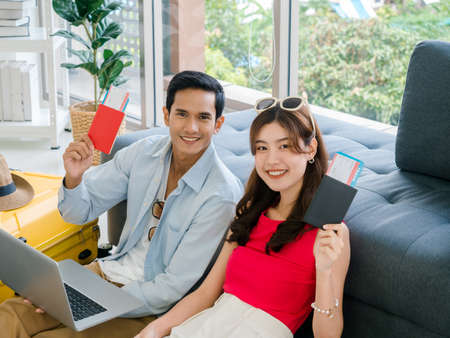 Happy Asian couple ready to trip, young man and woman, holding and showing passport while using laptop computer together for booking flight ticket, transport and hotel in living room, summer holiday.の写真素材