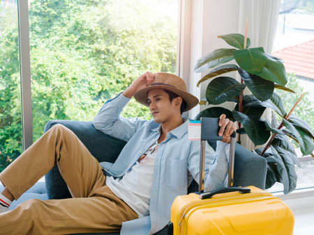 A man with yellow luggage. Handsome Asian male in denim shirt sitting and waiting alone while holding passport and yellow suitcase near the glass window. Summer holiday travel vacation concept.の写真素材