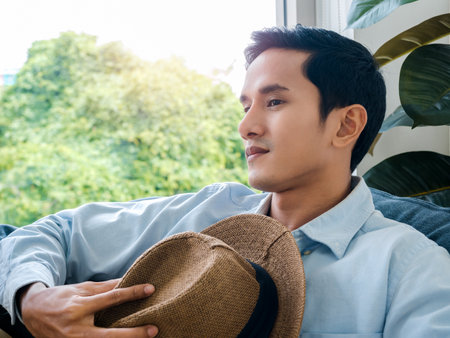 Portrait of handsome Asian man in light blue denim shirt and white tee holding brown beach hat sitting alone on cozy sofa seat near glass window and green leaves in the living room.の写真素材