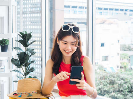 Young beautiful smiling Asian woman holding and using smart mobile phone, looking at screen. Happy Asian female wearing pink sleeveless, sunglasses, searching and planning for travel summer trip.の写真素材