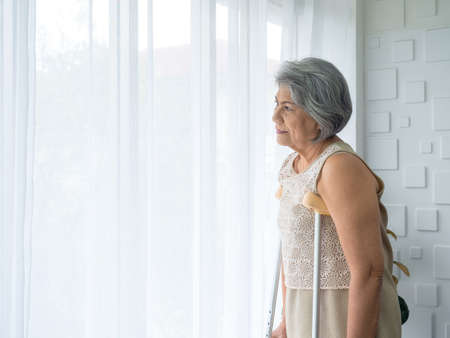Asian senior woman white hair standing with walking crutches near curtain looking out glass window with copy space. Elderly lady patient using crutches walker. Strong health, medical care concepts.の写真素材
