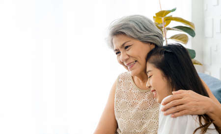 Portrait of happy Asian senior, mother white hair embracing her beautiful daughter with love, care, comfort and smile while sit on sofa on white background in living room at home with copy space.の写真素材