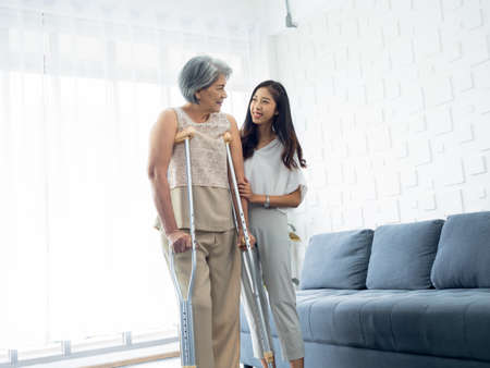 Young smiling Asian females carefully hold and support the arms of elderly women trying to walk on crutches in recovery room, helping old women, health care, therapy senior patient at home concept.の写真素材