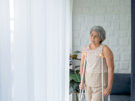 Asian senior woman white hair standing with walking crutches near curtain looking out glass window with copy space. Elderly lady patient using crutches walker. Strong health, medical care concepts.の写真素材