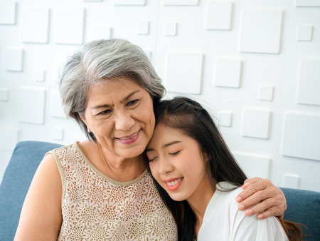Portrait of happy Asian senior, mother white hair embracing her beautiful daughter with closing eyes with love, care, comfort and smile while sit on sofa on white background in living room at home.の写真素材