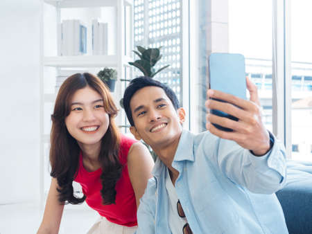 Happy handsome Asian man and young beautiful woman selfie while sitting near the glass window at...