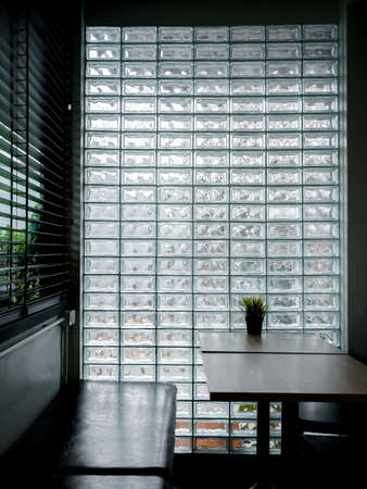 Glass brick wall background. Modern glass block wall with the light through decorated in the cafe with empty table and long seat, dim light room, vertical style. Interior decor space wall concept.の写真素材