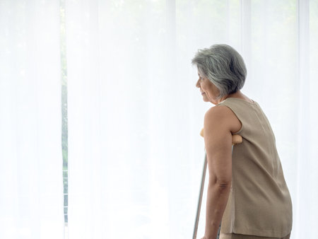 Asian senior woman white hair standing with walking crutches near curtain looking out glass window with copy space. Elderly lady patient using crutches walker. Strong health, medical care concepts.の写真素材