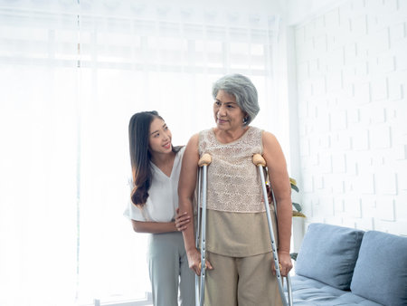 Elderly woman trying to walk on crutches standing held and supported in arms by young Asian female carefully in recovery room, helping old women, health care, senior therapy patient at home concept.の写真素材