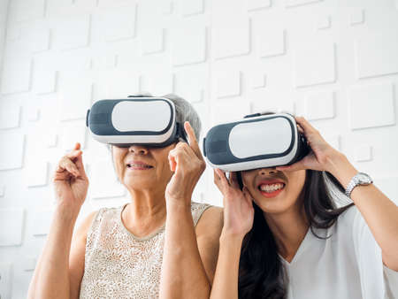 Asian young woman, happy daughter and her mother, old senior female wearing VR glasses, enjoy 3d game online together in white room. Mom and daughter with reality virtual technology lifestyle concept.の写真素材