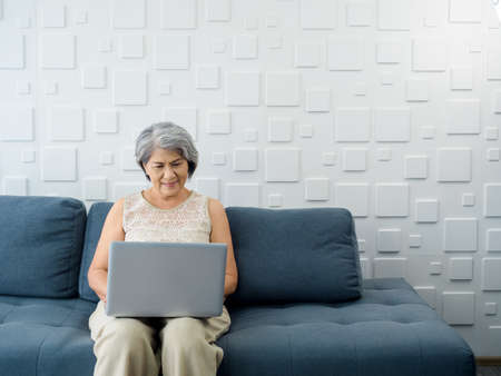 Portrait of smiling Asian senior casual woman sitting on the sofa, working with laptop computer in bright white room. Elderly female surf internet or video call at home. Older people with technology.の写真素材