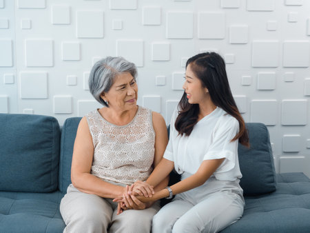 Portrait of happy Asian senior, mother or grandparent white hair holding hands of her beautiful daughter or grandchild smiling with love, care and comfort while sit on grey couch in living room.の写真素材