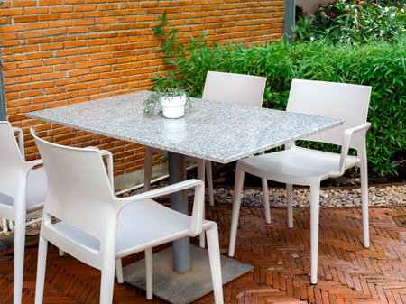 Four empty white plastic chairs with terrazzo table with small plant pot in the outdoor garden near the brick ground and wall building. Table set relaxing in garden.の写真素材