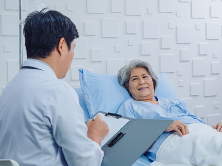 Happy and smiling Asian senior, old female patient lying on bed with receiving saline solution discussing with male doctor while he taking notes in hospital room. Health care and medical concept.の写真素材