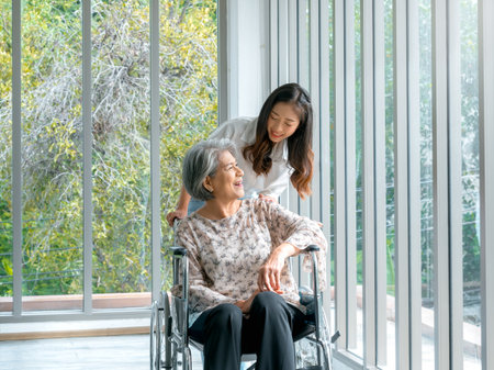 Happy Asian elderly woman, mother or grandparents on wheelchair taking care by caregiver, smiling young female, daughter or grandchild supporting at home on green nature background, senior healthcare.の写真素材