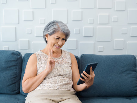 Happy Asian senior woman sit on couch, doing victory sign by hand at digital tablet screen in hand in white room. Elderly female meet with family by video call at home. Older people with technology.の写真素材