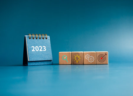 Business action plan with goal,  management, idea and research icon signs on wooden cube blocks with small blue desk calendar year 2023 on blue background. Business strategy concept, minimal style.の写真素材