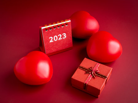 2023 year desk calendar and small red present gift box with heart balls on red background. An...