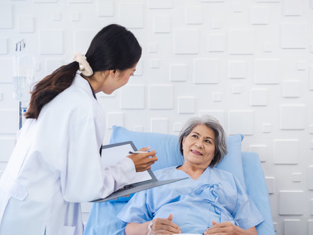 Smile Asian senior woman grey hair patient lying on bed, recovering while young female doctors visit, explaining the symptoms and examination notes and medical healthcare documents in hospital room.の写真素材