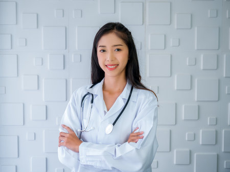 Smiling pretty Asian woman doctor portrait long hair, standing with arms cross in white medical office at hospital. Confident Asian young female practitioner with stethoscope, looking at camera.の写真素材