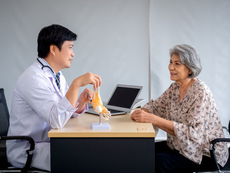 Smiling professional Asian man orthopedic doctor pointing to foot skeleton, ankle joint anatomy model to explain for senior female patient in medical office. Physio treatment, health care concept.の写真素材