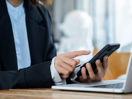 Pointing finger of businesswoman touching on cellphone. Black smart mobile phone in person hand holding near the laptop computer on desk. Cyber security, investment and business technology concepts.の写真素材