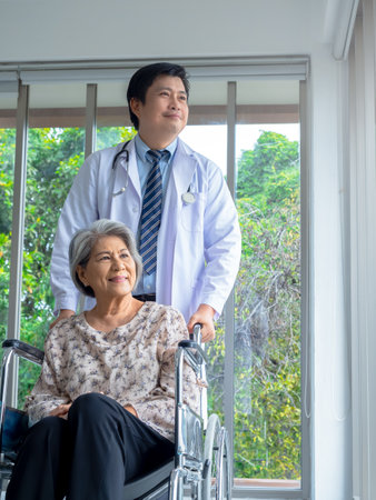 Smiling happy Asian elderly senior female patient sits in wheelchair with man doctor in white coat standing in medical office in hospital, vertical style. Caregiving, healthcare and medical concept.の写真素材