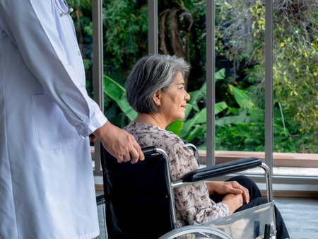 Close-up portrait of happy Asian elderly senior female patient sits in wheelchair while man doctor in white coat take care in medical office in hospital. Caregiving, healthcare and medical concept.の写真素材