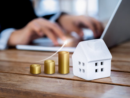 Property value, real estate investment, mortgage and home tax concepts. Rising arrow on golden coins stack as a graph near miniature house near businessman work with laptop computer on wooden table.の写真素材