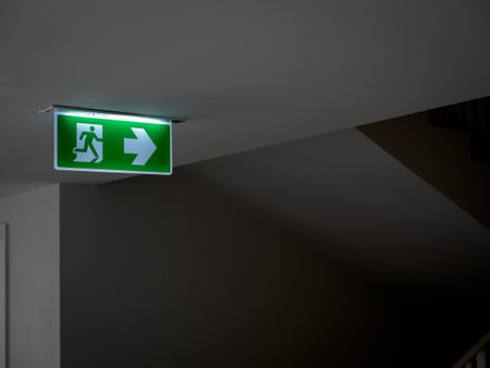 Green fire escape sign hang on the ceiling in the dark building near the stairway. Emergency fire exit sign, warning plate with running man icon and arrow to the right way with copy space.の写真素材